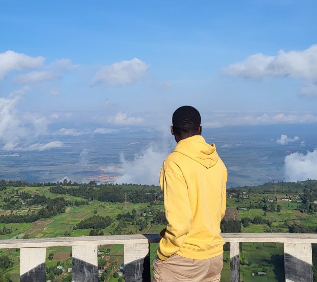Person i gul hoodie står vid ett räcke och blickar ut över ett grönt landskap och en vid horisont under blå himmel.